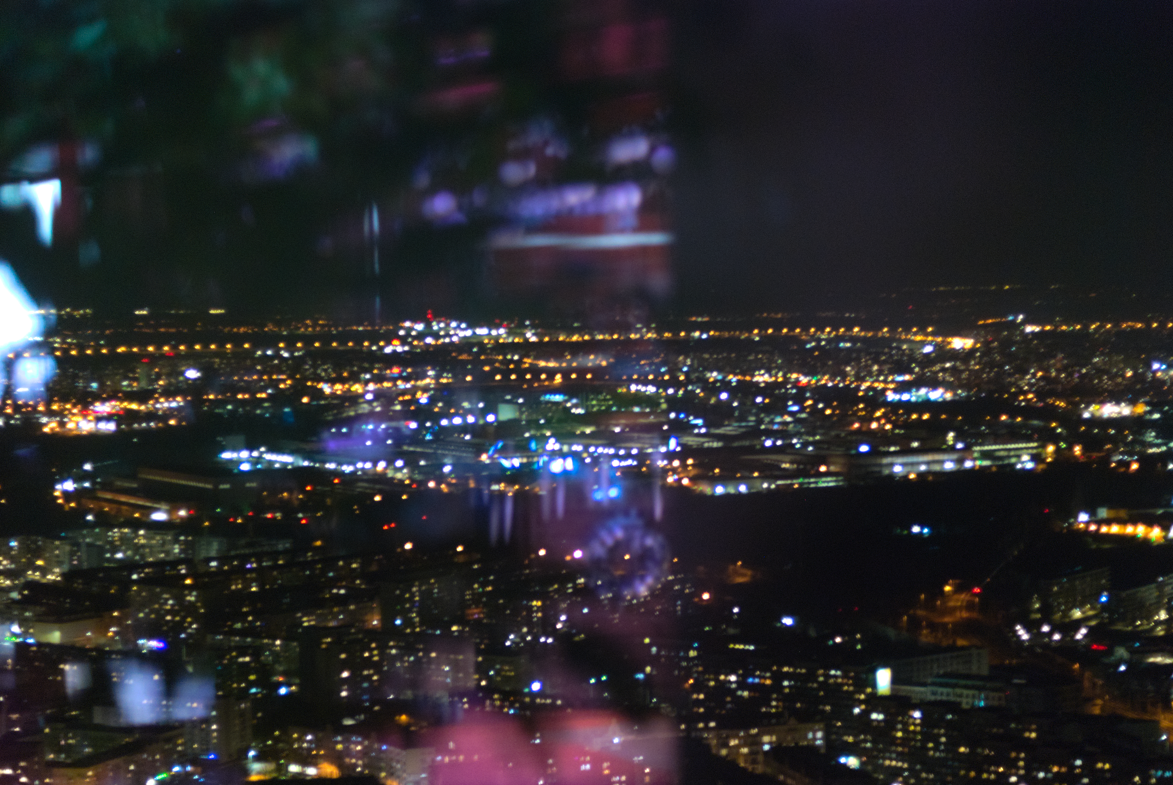 wide angle shot of a city at night, captured from very high up (approximately the 50th floor). the city's night life and residential lighting provide a rough outline of the buildings otherwise shrouded in the darkness of a cold winter night. an indecipherable reflection suggests the photo was taken from behind a window.