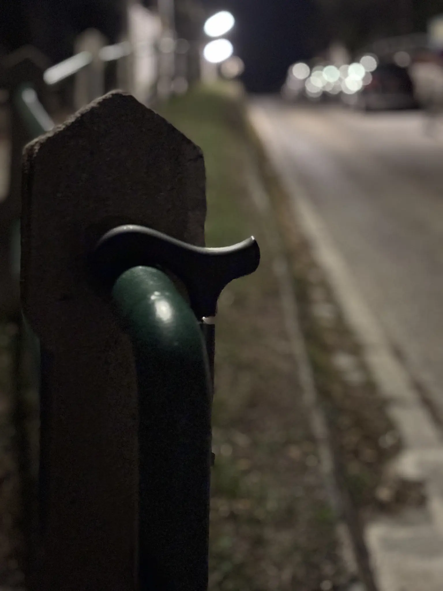 a cane made from carbon fibre, a metal ring and a rubber handle has its handle placed on a thick metal railing that has been painted with a dark green. the metal rail is being held up by concrete pillars with   pyramid-shaped tops. a street with a few parked cars and street lightning can be seen in the blurry background. it seems to lead up a hill; the railing has been built next to a pathway that is separated from the small street with a narrow grassy patch.