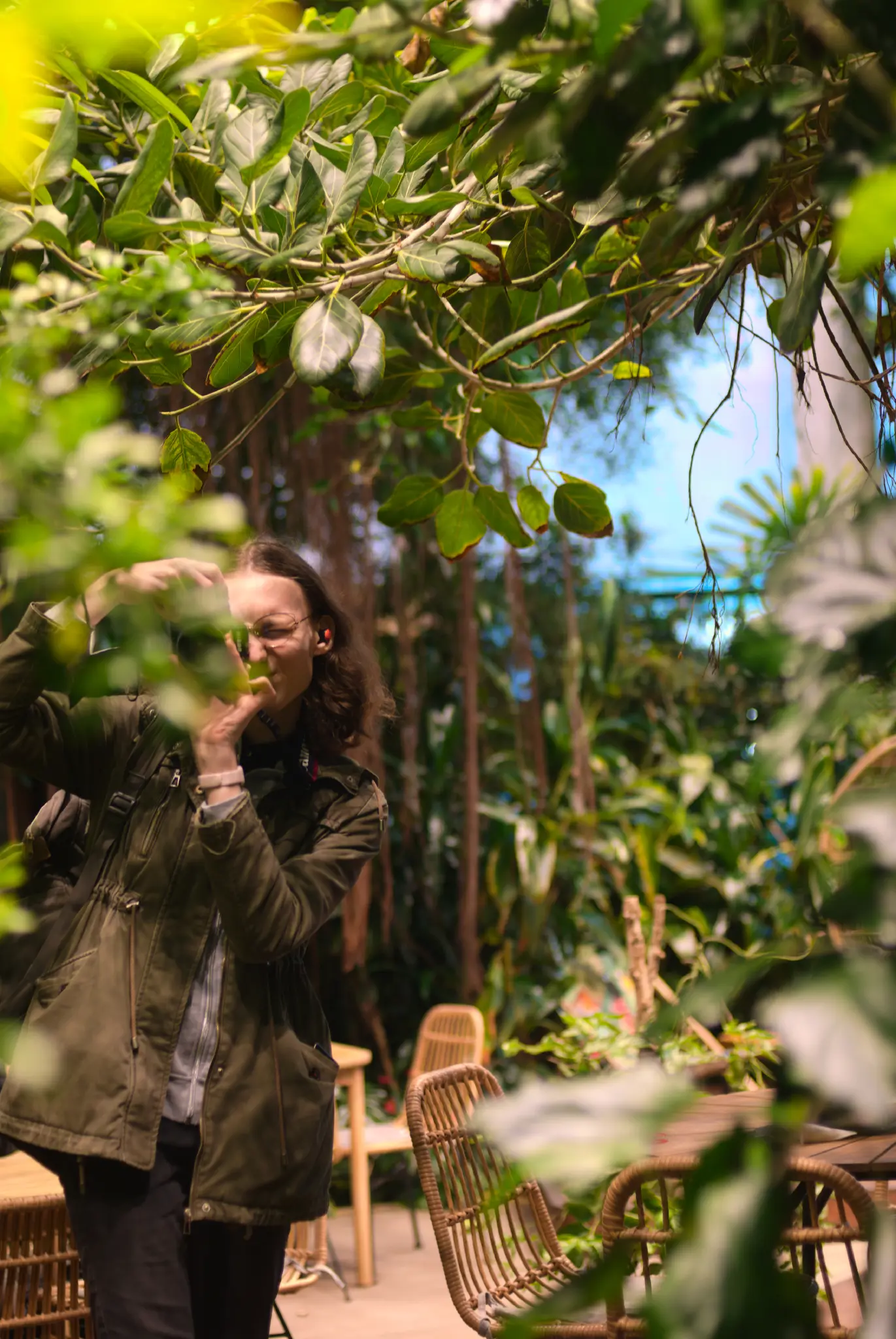 a white woman with shoulder length curly hair surrounded by trees in an indoor tropical garden, with branches with oval-shaped leaves framing and slightly obscuring her face. she's looking into a camera viewfinder, and adjusting focus on the lens. she's wearing a khaki parka over a grey zipper hoodie, and dark grey pants. she's standing next to some wooden tables and chairs.