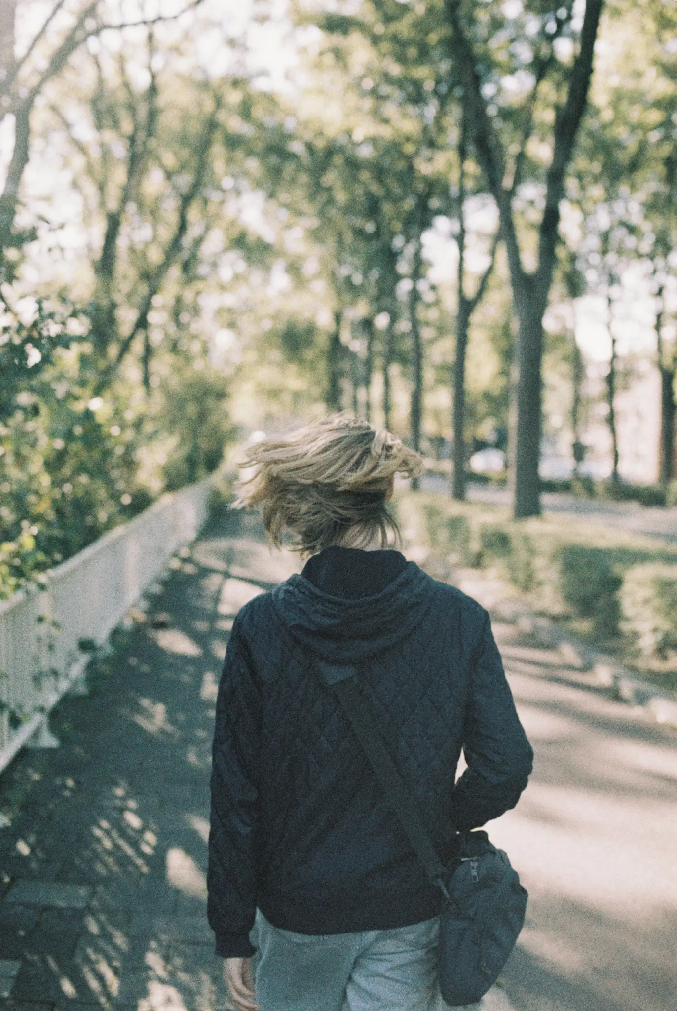 person swirling their chin-length hair while walking down a pedestrian walkway with trees on either side. they're facing away from the camera, and are wearing a dark navy blue jacket, light blue jeans and a gray messenger bag. their left hand is dangling, while they're using their right hand to hold the messenger bag in place.
the harsh sunlight shining through the trees is blowing out the leaves on smaller branches, giving the whole scene somewhat of an ethereal vibe.