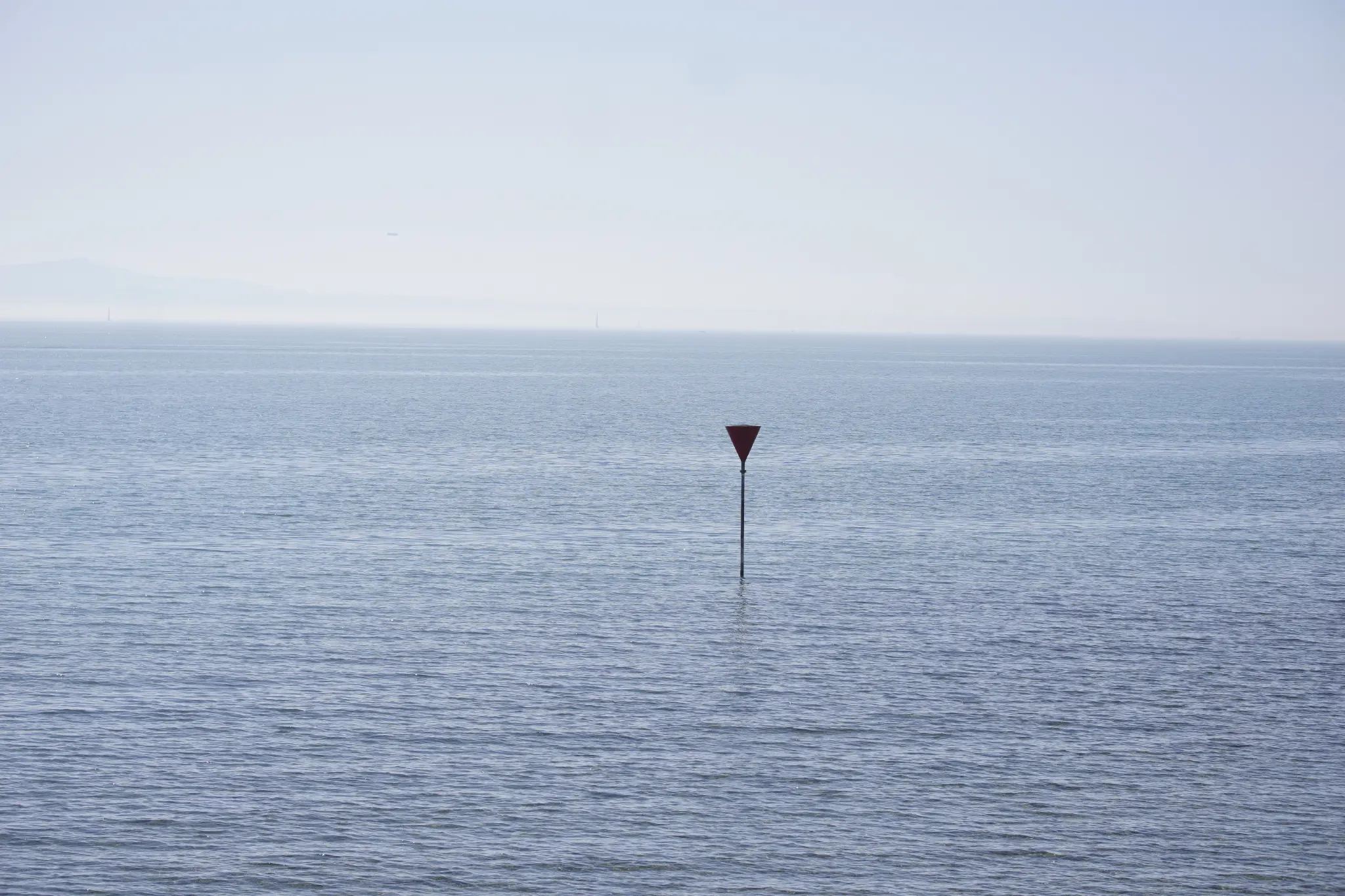 a singular red triangular indicator stands out from a slightly choppy lake constance. it’s quite a misty day, meaning that there is only a very faint outline of a few mountains in the horizon.