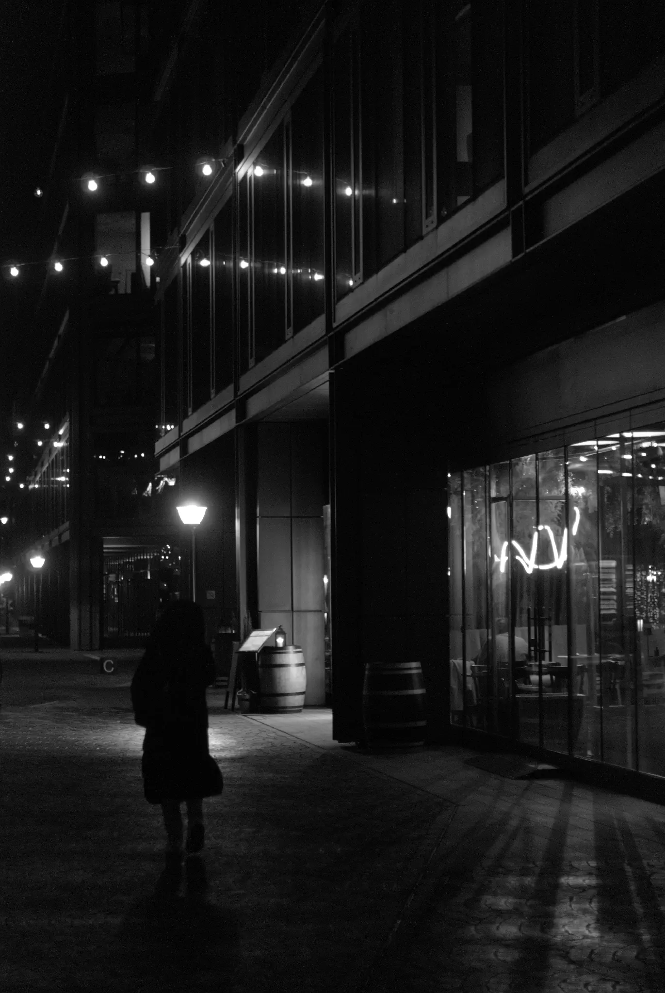 black and white photo of a person in a coat walking in a dimly lit pedestrian space, with neon lights in a restaurant casting harsh, long shadows of the tall frames supporting the store's glass front. lightbulbs strung up on a cable and streetlights are providing a soft background lighting. the lightbulbs are reflecting in the glass facade on the first floor above the restaurant, creating a satisfying arc shape.