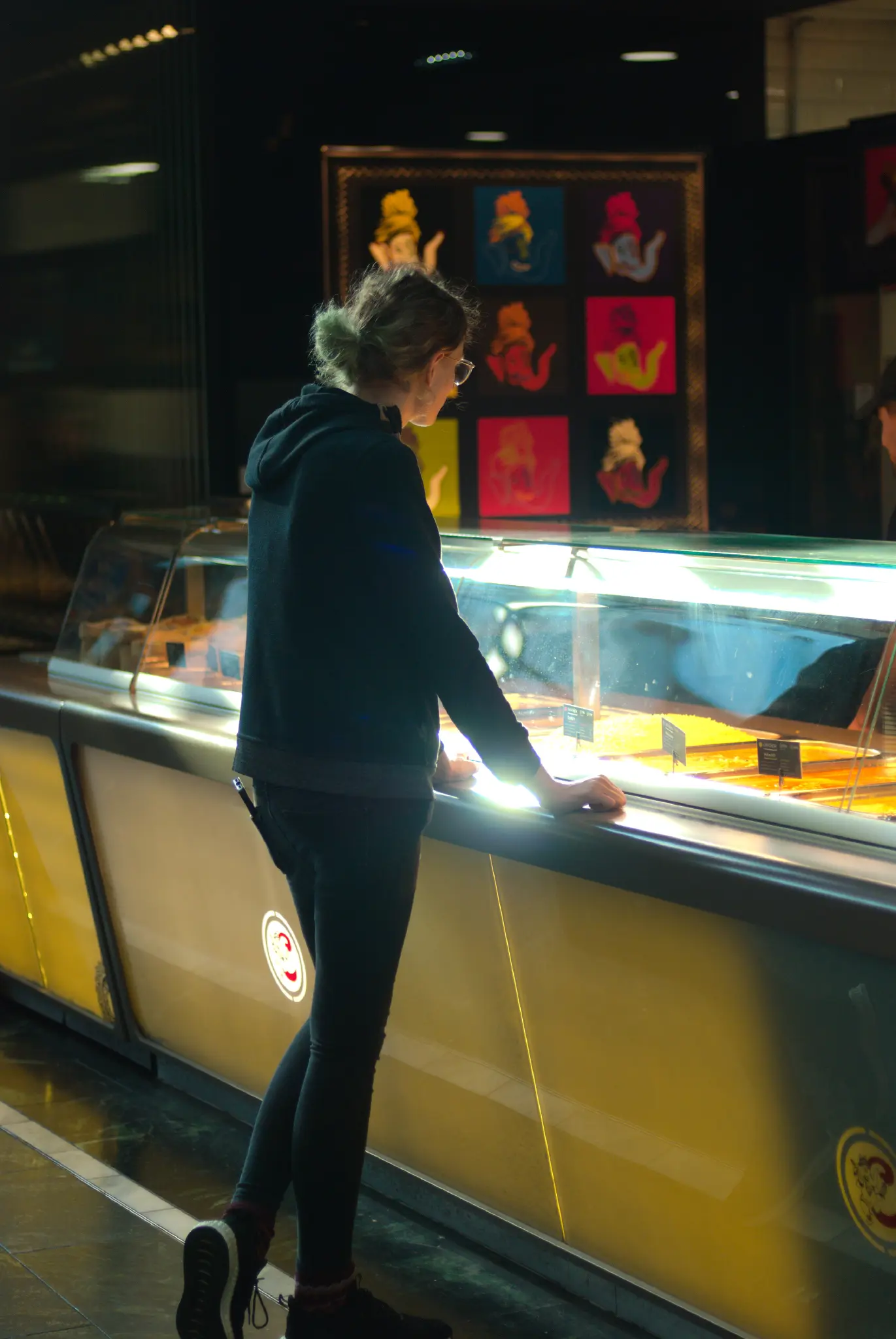 a person standing at the counter at an indian takeout restaurant. their hands are resting on the countertop, and the sun is creating a blindingly bright reflection between them. the person's left foot is angled upwards, with the tip of their shoe resting on the floor. a row of orange to yellow foods are visible on the counter behind a glass cover. in the background, some artwork is hung up on a wall.
