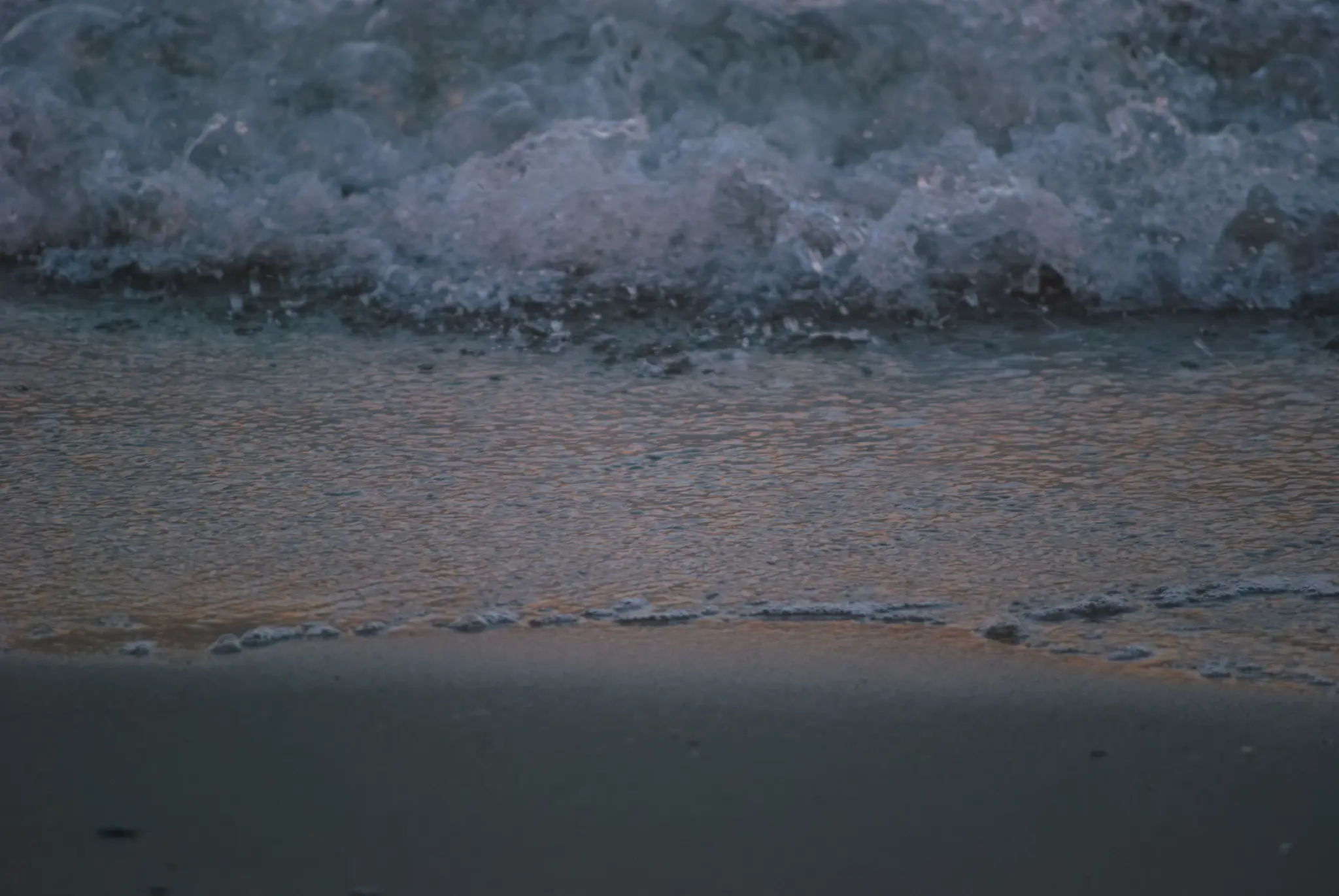 a wave crashing onto a beach, with the sunset reflecting in the much calmer remains of the previous wave flowing back into the ocean. slightly blurry droplets of sea foam and water are launched a few centimeters off the surface.