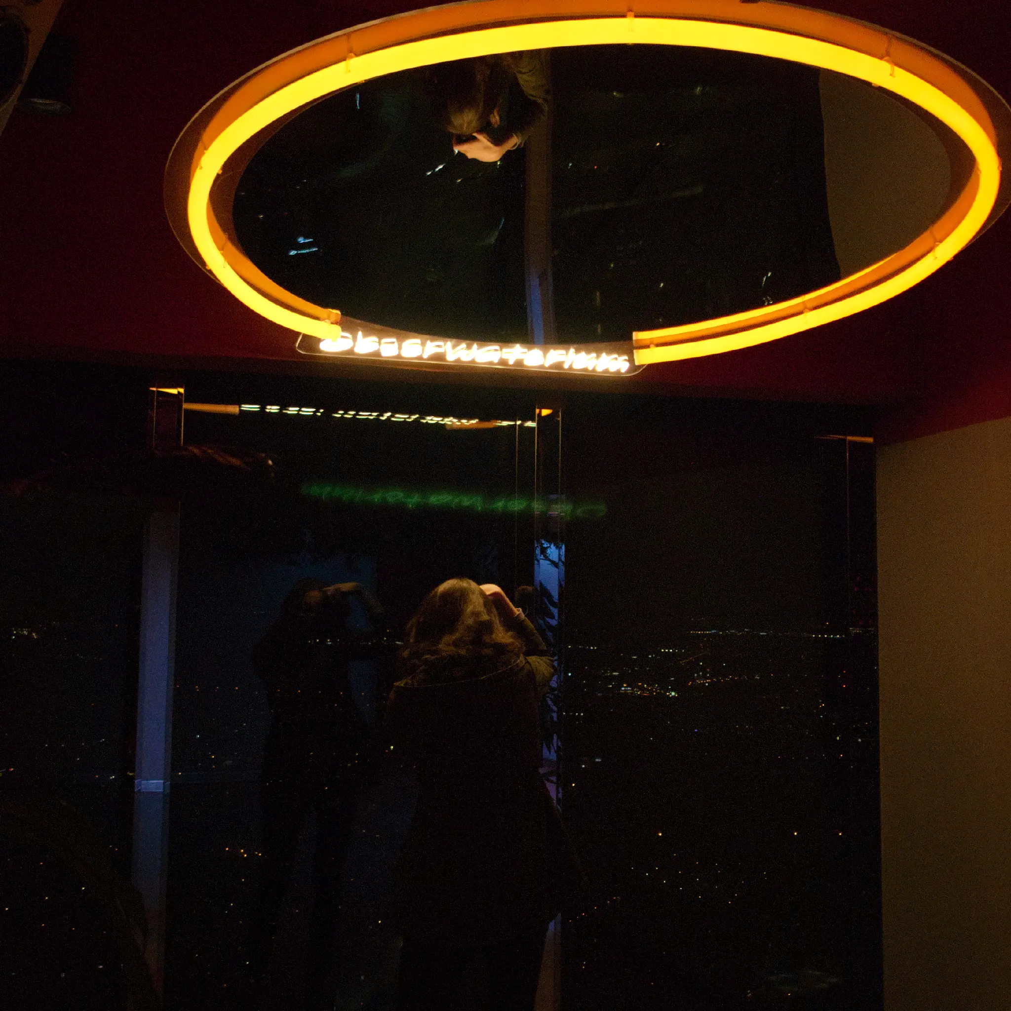 a young white woman with brown shoulder length hair standing at a window in a skyscraper, facing outside. she's looking into her camera's viewfinder, wearing a khaki parka and dark grey pants. there's some neon text above her, reading "obserwatorium", and next to it a large round mirror is mounted from the ceiling. her reflection is visible in it, as well as in the window she's photographing out of.