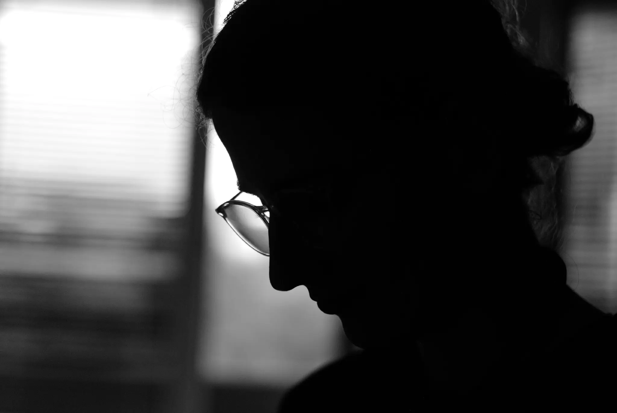 a black and white image showing the black silhouette of a person's head and shoulders in front of a window. the person is wearing glasses, has tied up curly hair, and the outline of their nose, mouth and chin are visible.