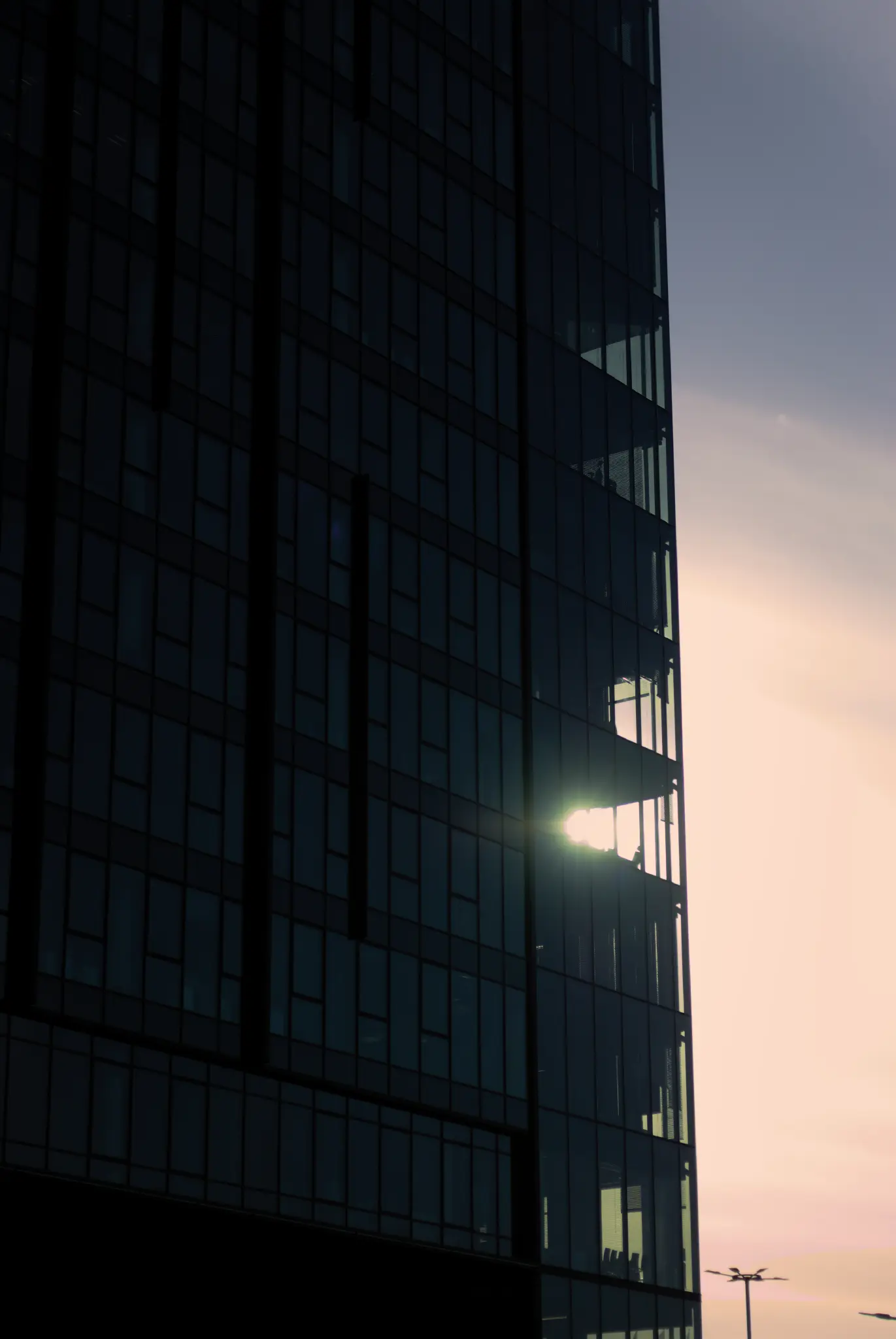 a high-rise office building with the sun shining through its corner. the sky is illuminated in gentle pastel hues ranging from a pink-ish orange near the horizon to a slightly darker blue towards the sky. the building itself is pictured very dark, creating a harsh contrast between it and the well-lit sky.