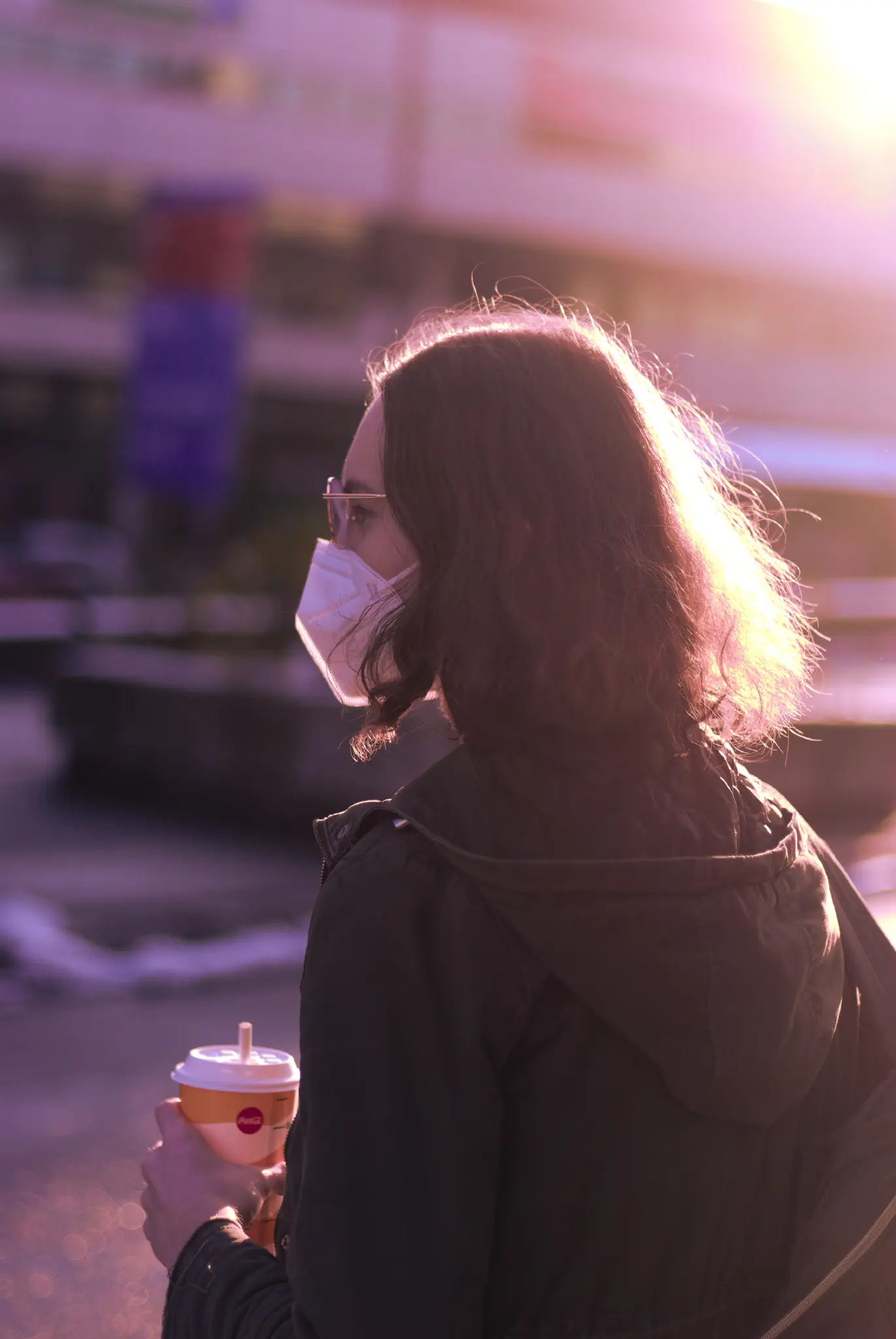 a young white woman with neck-long brown curly hair wearing a khaki parka, glasses and a white ffp2 mask. she's carrying a backpack and a cup with a paper straw. the afternoon sun is illuminating the back of her hair, giving it an unusual golden glow that fades into a pink-ish purple. the whole image is color graded that way: highlights are a golden or yellow color, and shadows lean towards purple.