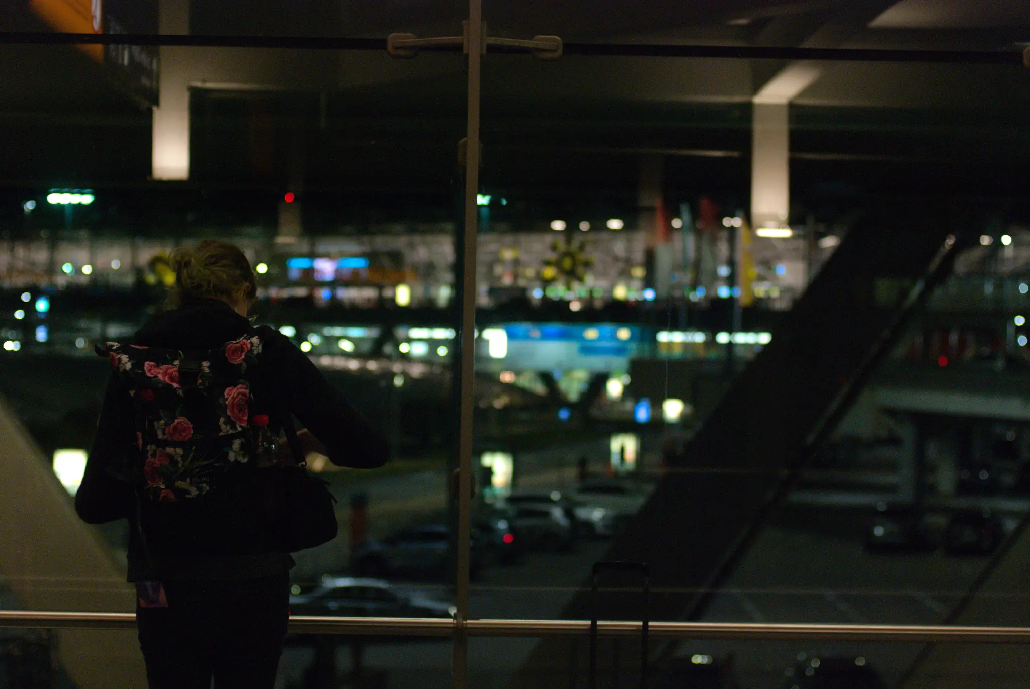 a person with tied up hair in black clothing and a backpack with flowery print standing at a window overlooking a parking lot and an airport terminal. they are looking down in front of themselves, and there's a phone with a colorful case in their back pocket.